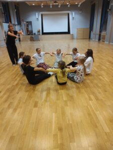 Lapsia istumassa piirissä lattialla käsistä kiinni pitäen. Ohjaaja seisoo ringin ulkopuolella.