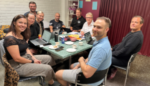 Miehiä ja naisia istuu pöydän ympärillä ja katsoo kameraan.