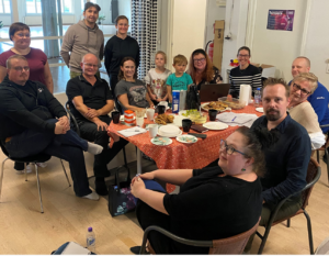 Ryhmäkuva joss pöydän ääressä istuu miehiä ja naisia, jotka katsovat kameraan.